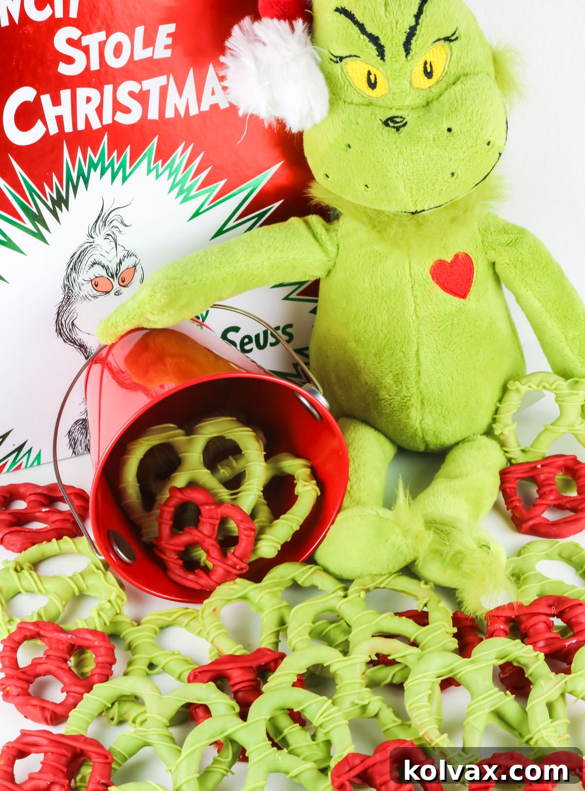 Grinch Heart Pretzels spilling out of a red metal pain onto a white surface surrounded by a Grinch stuffed animal and a How the Grinch Stole Christmas book.