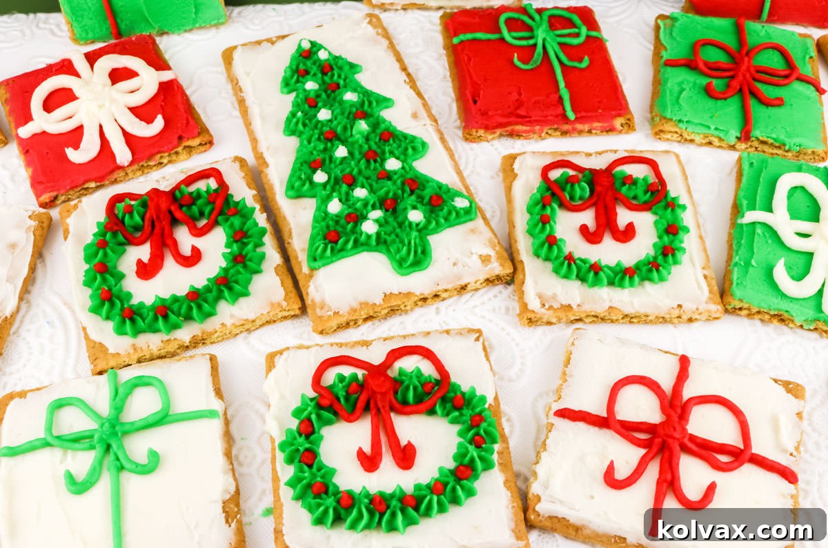 A beautiful batch of Christmas Frosted Graham Crackers laying on a white table, adorned with festive designs like green wreaths with red berries and sparkling Christmas trees.