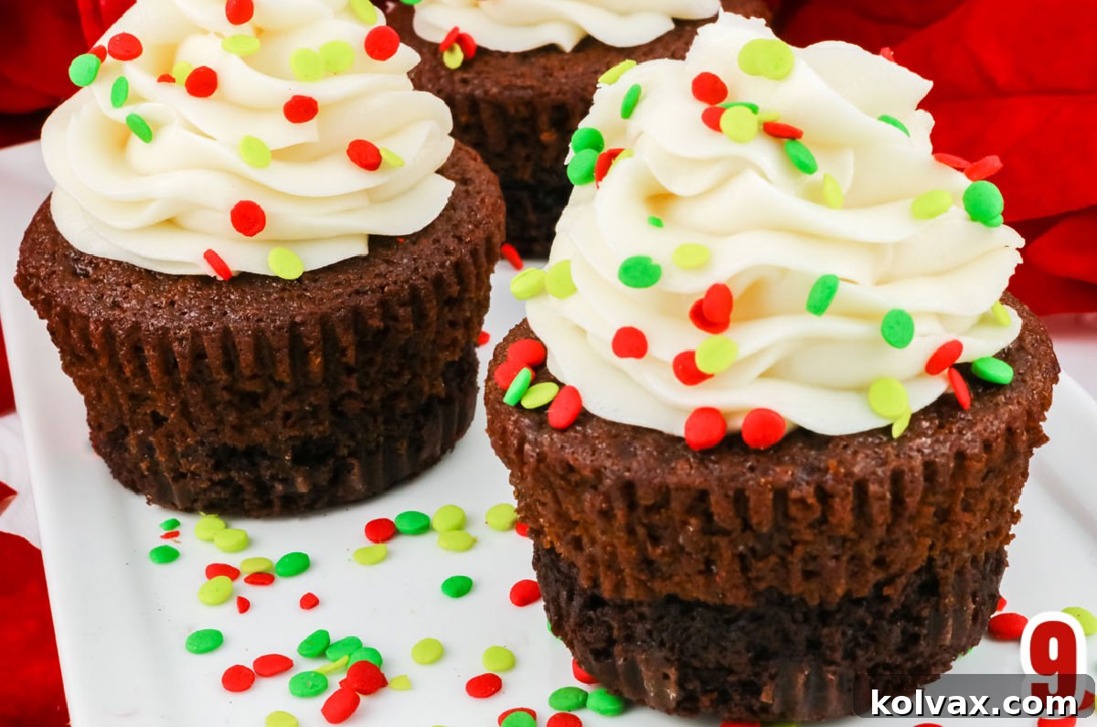 Three beautifully decorated Brownie Gingerbread Cupcakes, frosted with elegant swirls of white buttercream and adorned with colorful Christmas sprinkles, arranged on a white serving platter against a festive red background.