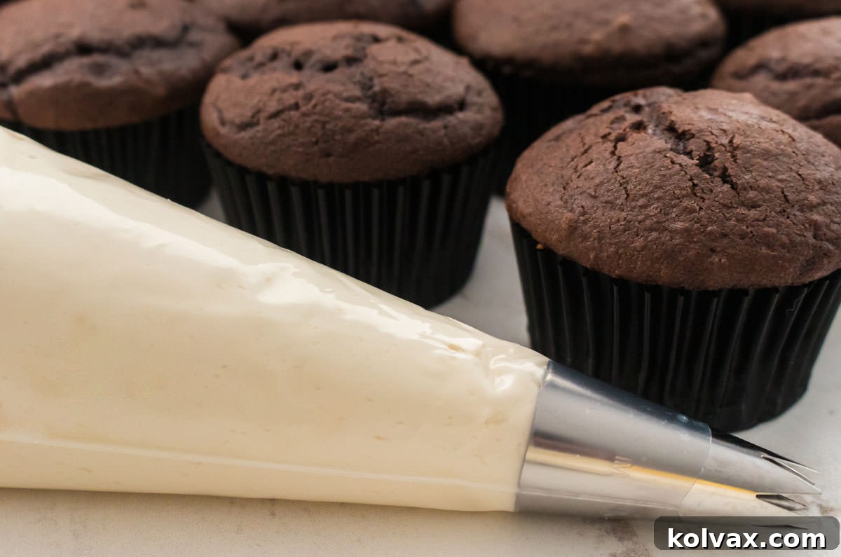 A pastry bag filled with smooth Baileys Irish Cream Frosting, ready for decorating, positioned in front of a batch of unfrosted chocolate cupcakes.