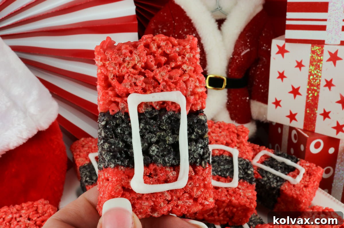 A hand holding up a single Santa Claus Rice Krispie Treat, showcasing its iconic red and black design, in front of a platter full of the remaining festive treats.