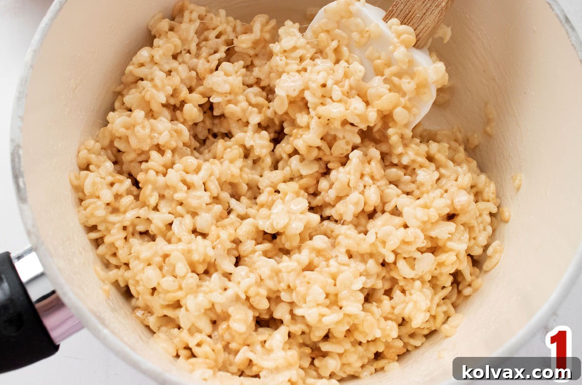 Closeup view of a white pan filled with freshly mixed, warm Rice Krispie Treat mixture and a white wooden spoon, ready for shaping.