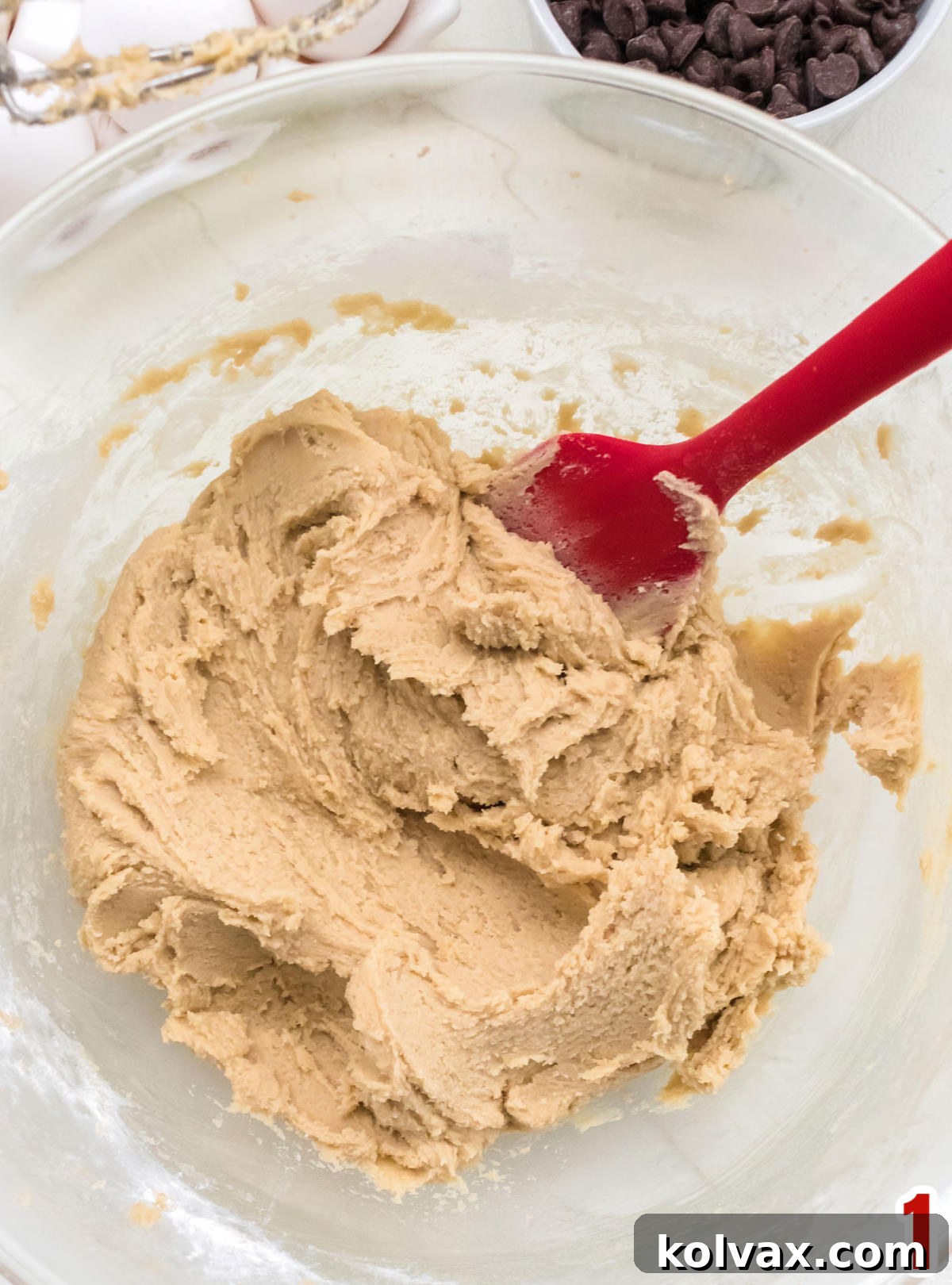 Overhead closeup on a clear glass bowl filled with chocolate chip cookie dough and a red spatula, ready for coloring.