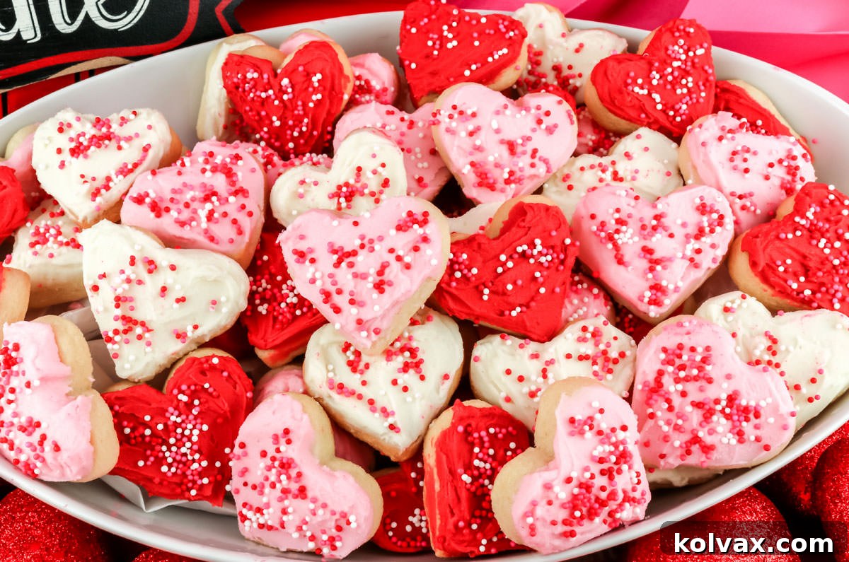 A white serving bowl brimming with Mini Heart-shaped Sugar Cookies, beautifully frosted in shades of pink, white, and red buttercream, and adorned with festive Valentine's sprinkles.