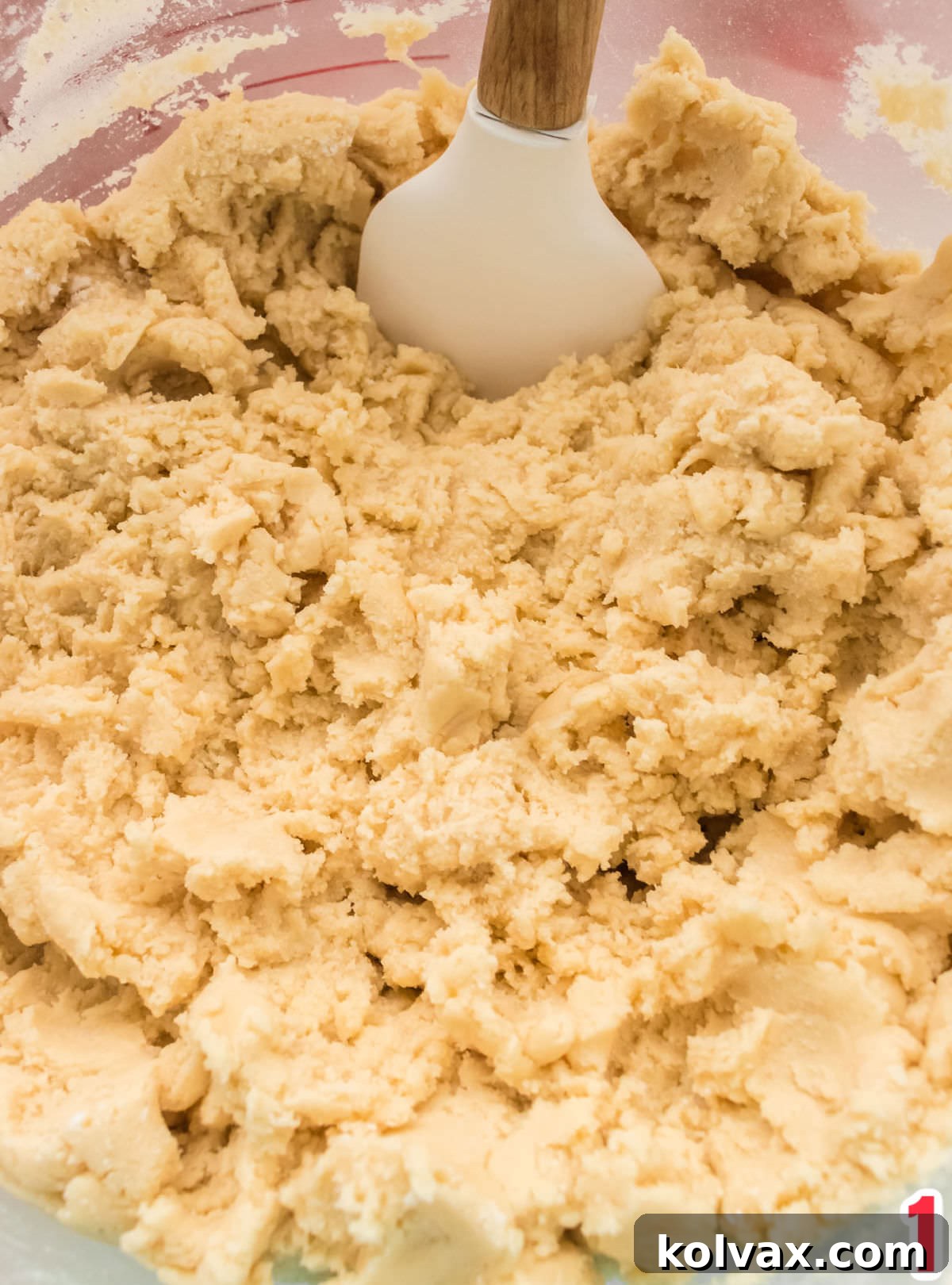 A close-up view of impeccably prepared sugar cookie dough resting in a clear glass bowl, accompanied by a wooden spatula, ready for rolling.