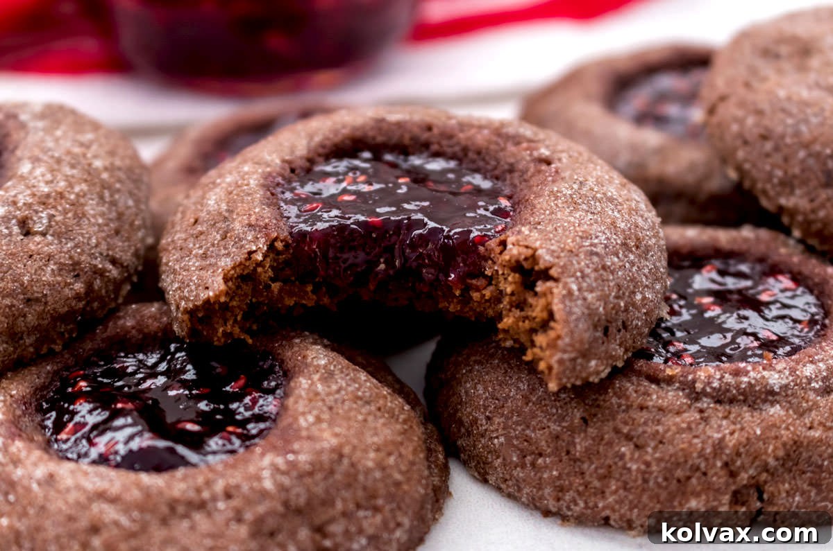 Close-up of a stack of rich Chocolate Thumbprint Cookies, with one cookie showing a bite taken out, revealing the vibrant red raspberry jam center.