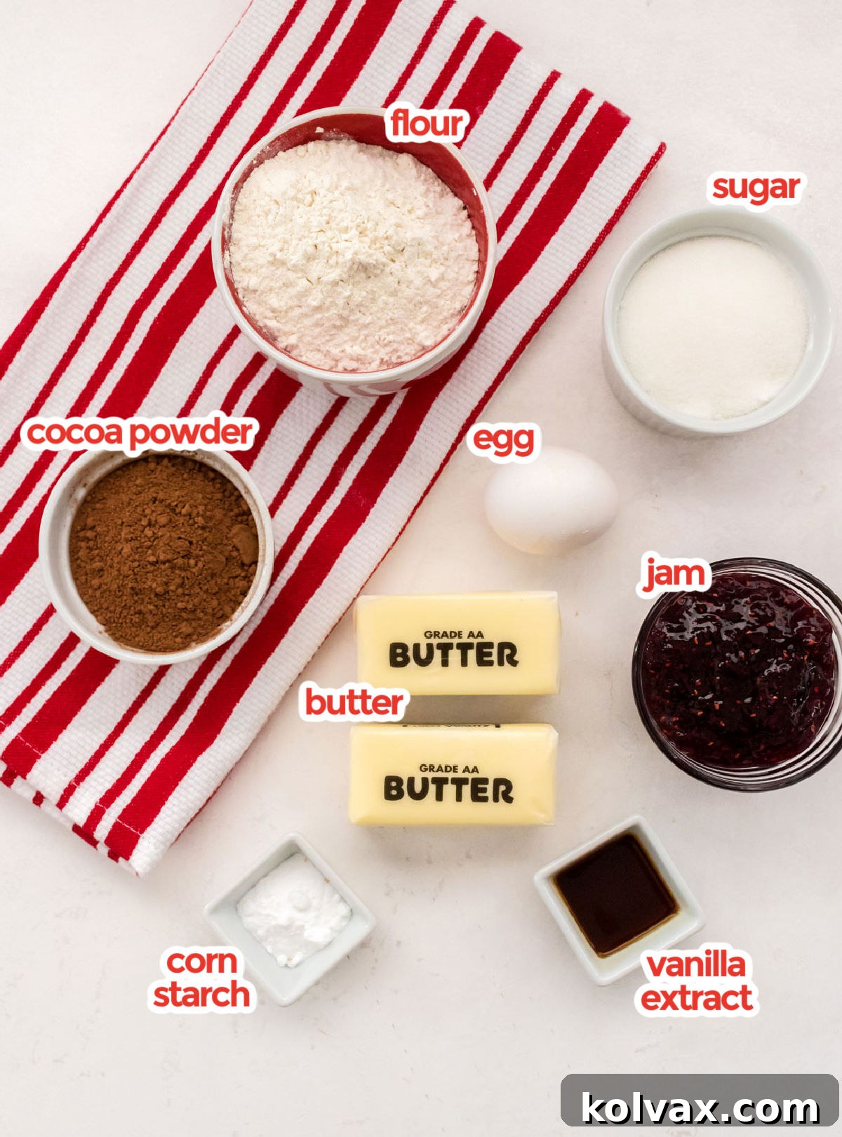 A flat lay of all the essential ingredients for making Chocolate Thumbprint Cookies, including flour, sugar, egg, cocoa powder, softened butter, red raspberry jam, vanilla extract, and cornstarch.