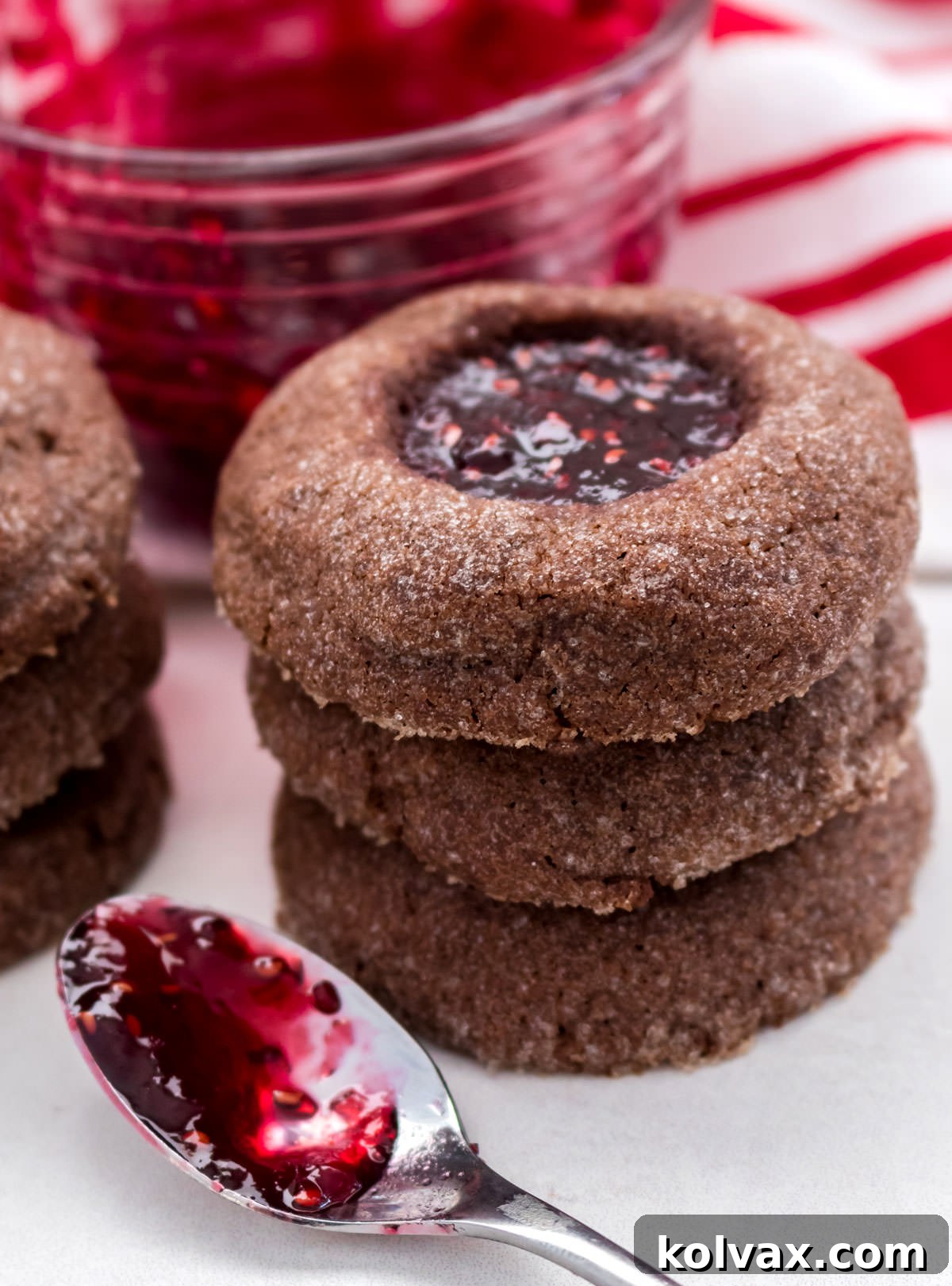 A close-up shot of a stack of rich Chocolate Thumbprint Cookies, with a glass bowl of Red Raspberry Jam and a jam-covered spoon blurred in the background, creating a mouth-watering visual.