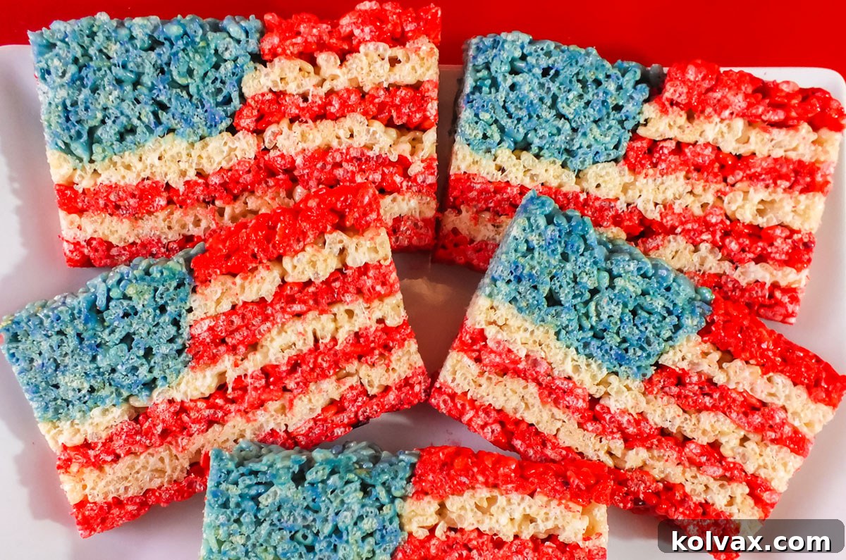 Closeup on a plate of vibrant Red, White, and Blue 4th of July Rice Krispie Treats, perfectly shaped like the American Flag, sitting on a white serving platter atop a festive red table cloth.
