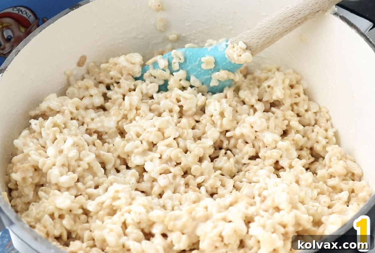 A white saucepan filled with a freshly mixed, gooey Rice Krispie Treat mixture, ready for shaping, accompanied by a blue wooden spatula.