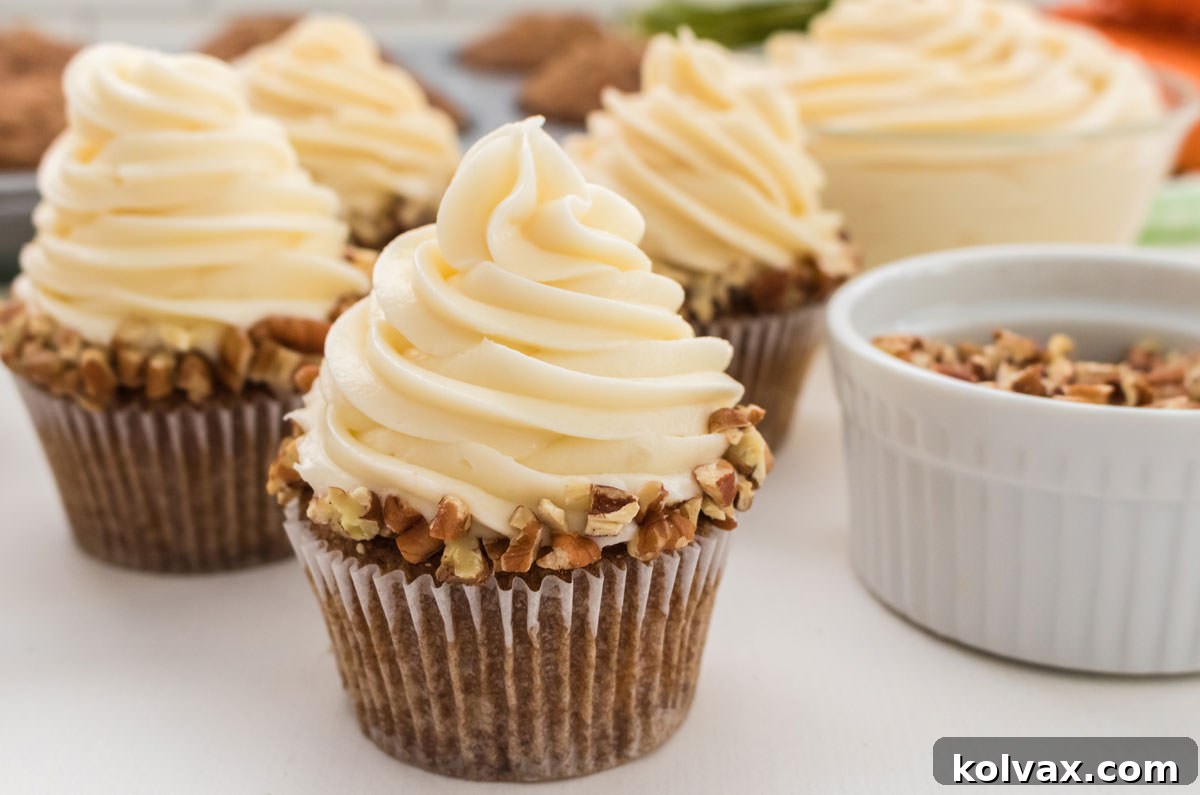 Four perfectly frosted Carrot Cake Cupcakes with Cream Cheese Frosting, adorned with chopped nuts, on a white table. In the background, a bowl of frosting and a cupcake tin hint at more deliciousness.