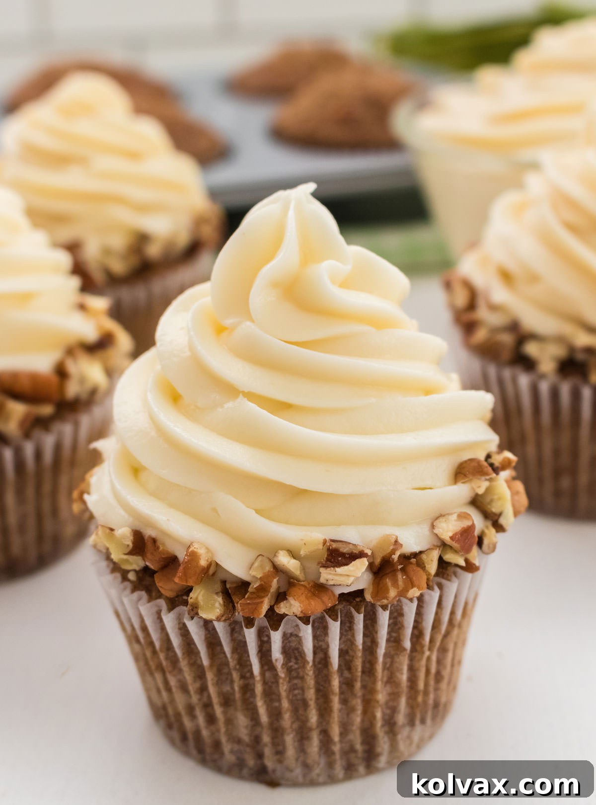 A collection of beautifully decorated homemade Carrot Cake Cupcakes on a white table, with unfrosted cupcakes in a tin in the background, showcasing the baking process.