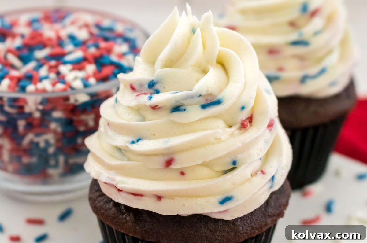 Two chocolate cupcakes generously frosted with Homemade Ice Cream Buttercream Frosting, adorned with colorful sprinkles, next to a bowl of extra sprinkles.