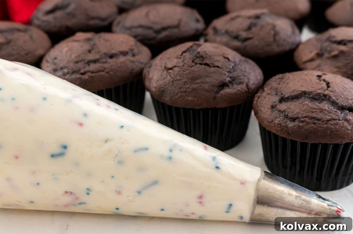 A decorating bag filled with luscious Homemade Ice Cream Buttercream Frosting, poised in front of a tray of unfrosted chocolate cupcakes.