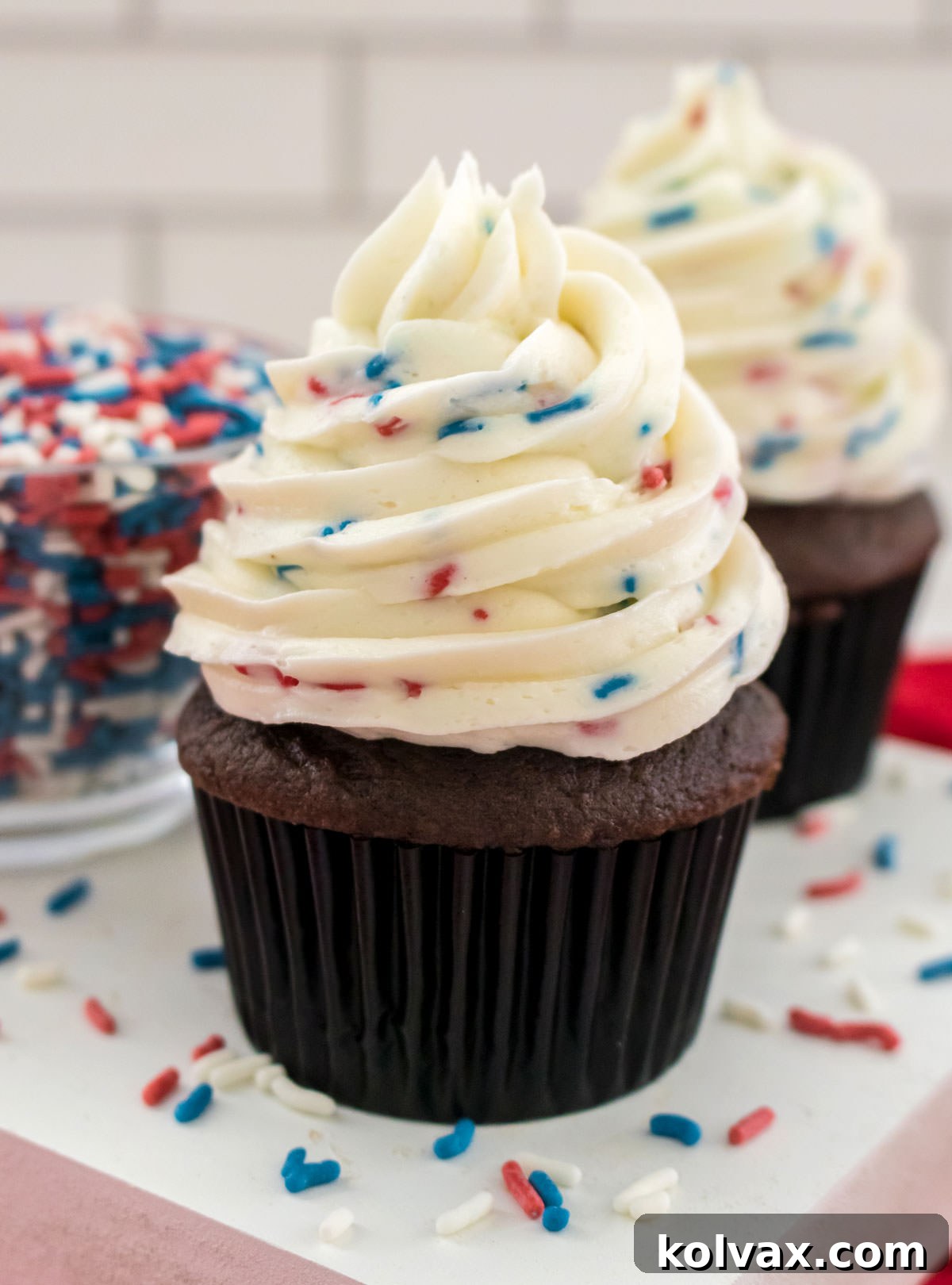 A beautifully frosted chocolate cupcake with Homemade Ice Cream Buttercream Frosting, garnished with sprinkles, resting on a white table next to a bowl of extra sprinkles.