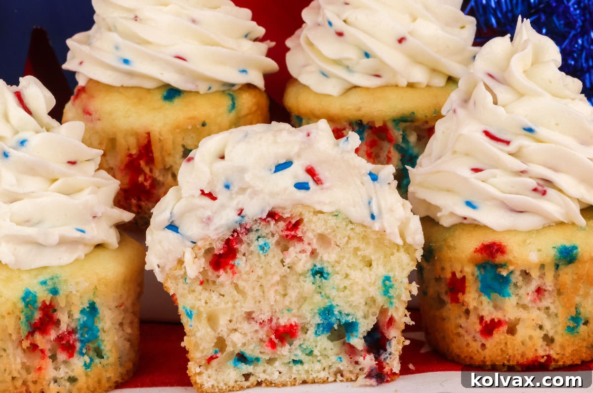 A striking close-up photograph showcasing five vibrant Red, White, and Blue Confetti Cupcakes resting on a crisp white table, with one cupcake expertly cut in half to reveal its cheerful sprinkle-filled interior.