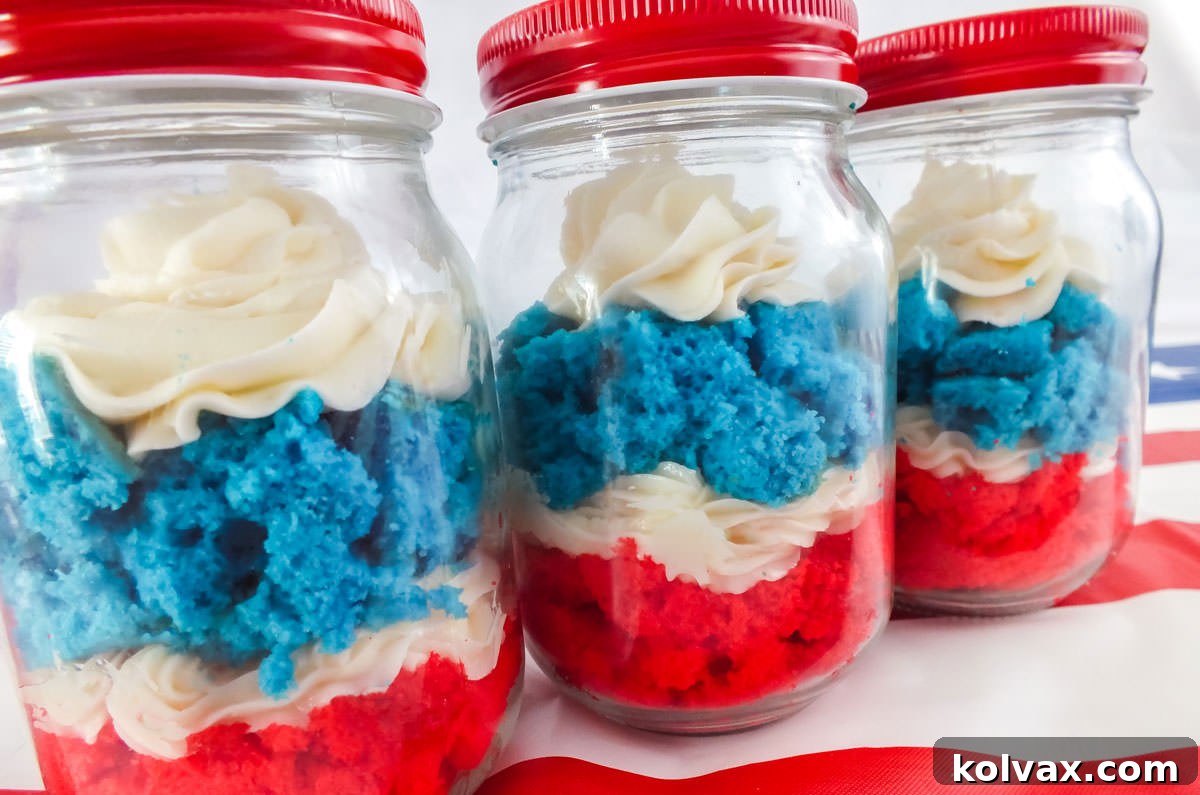 Three beautifully layered Red, White, and Blue Cupcakes in Jar, perfectly presented on a classic red striped tablecloth, set against a clean white background, ideal for picnics.