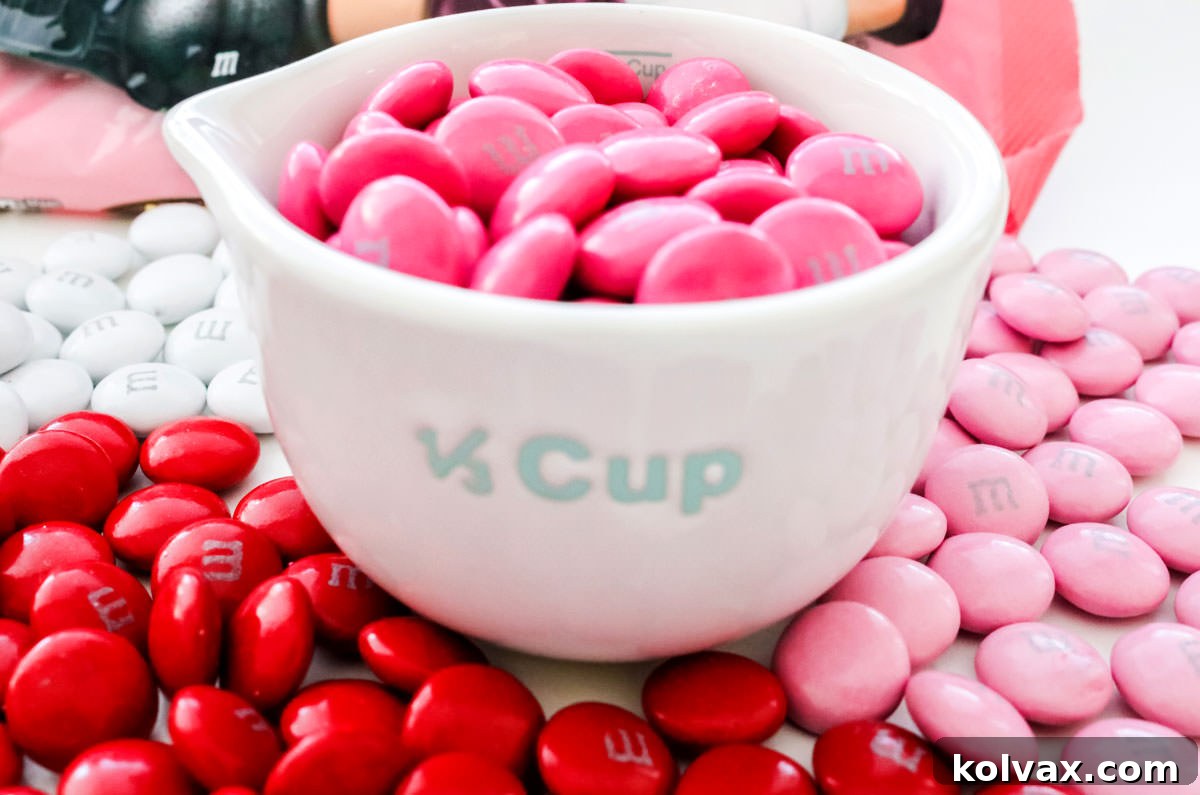 White measuring cup holding dark pink M&M's surrounded by red, light pink and white M&M's.