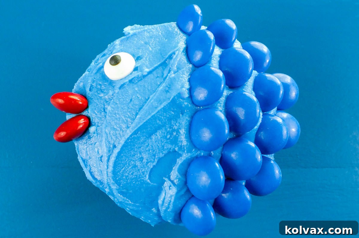 A vibrant blue Little Fishy Cupcake, adorned with colorful M&M scales and candy eyes, sitting on a matching blue surface, ready to charm.