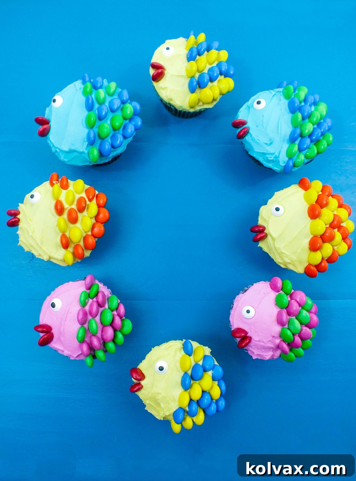 Eight cheerful Little Fishy Cupcakes, showcasing a variety of colors and designs, are arranged in a charming circle on a smooth blue surface.
