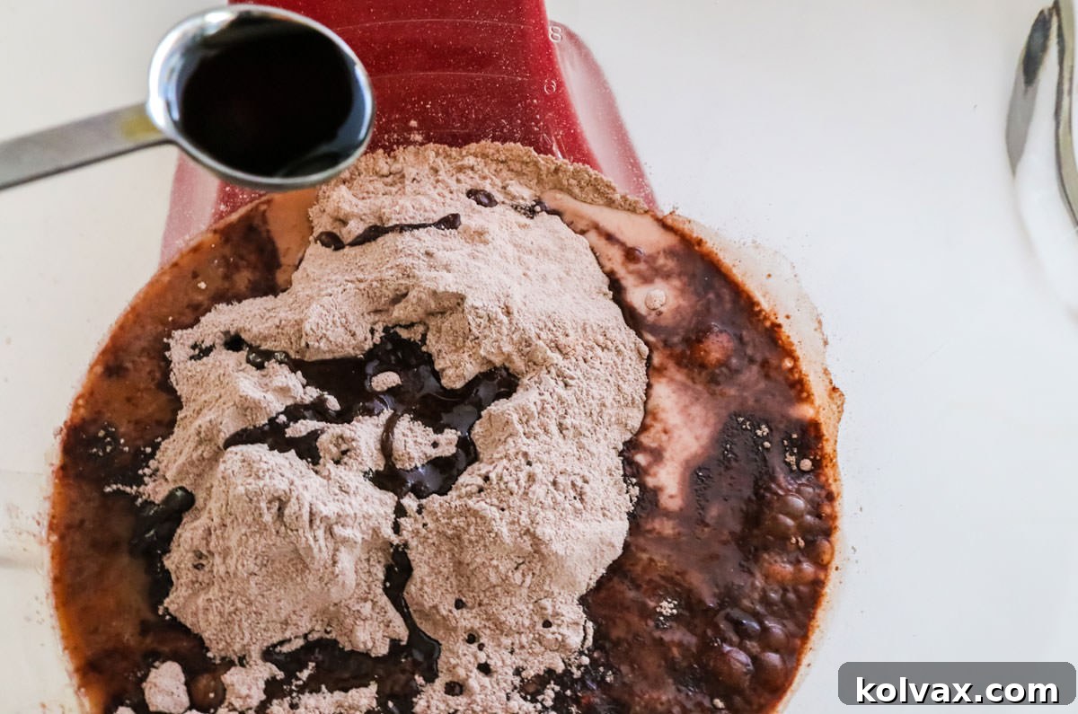 A small measuring spoon adding a teaspoon of pure vanilla extract into a clear mixing bowl filled with dark chocolate cake batter. The focus is on the liquid drop.