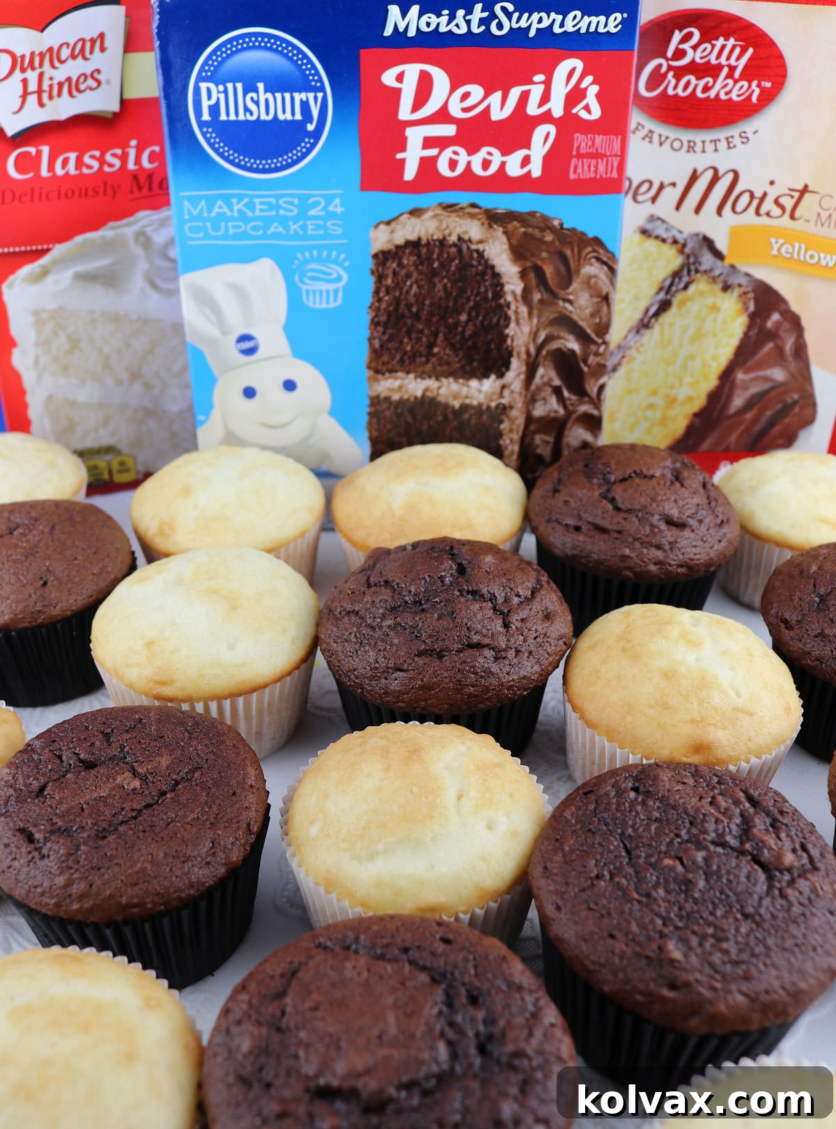 A vibrant display of unfrosted chocolate and vanilla cupcakes arranged on a white table, positioned in front of a selection of popular store-bought cake mix boxes. The scene captures the essence of baking with convenience.