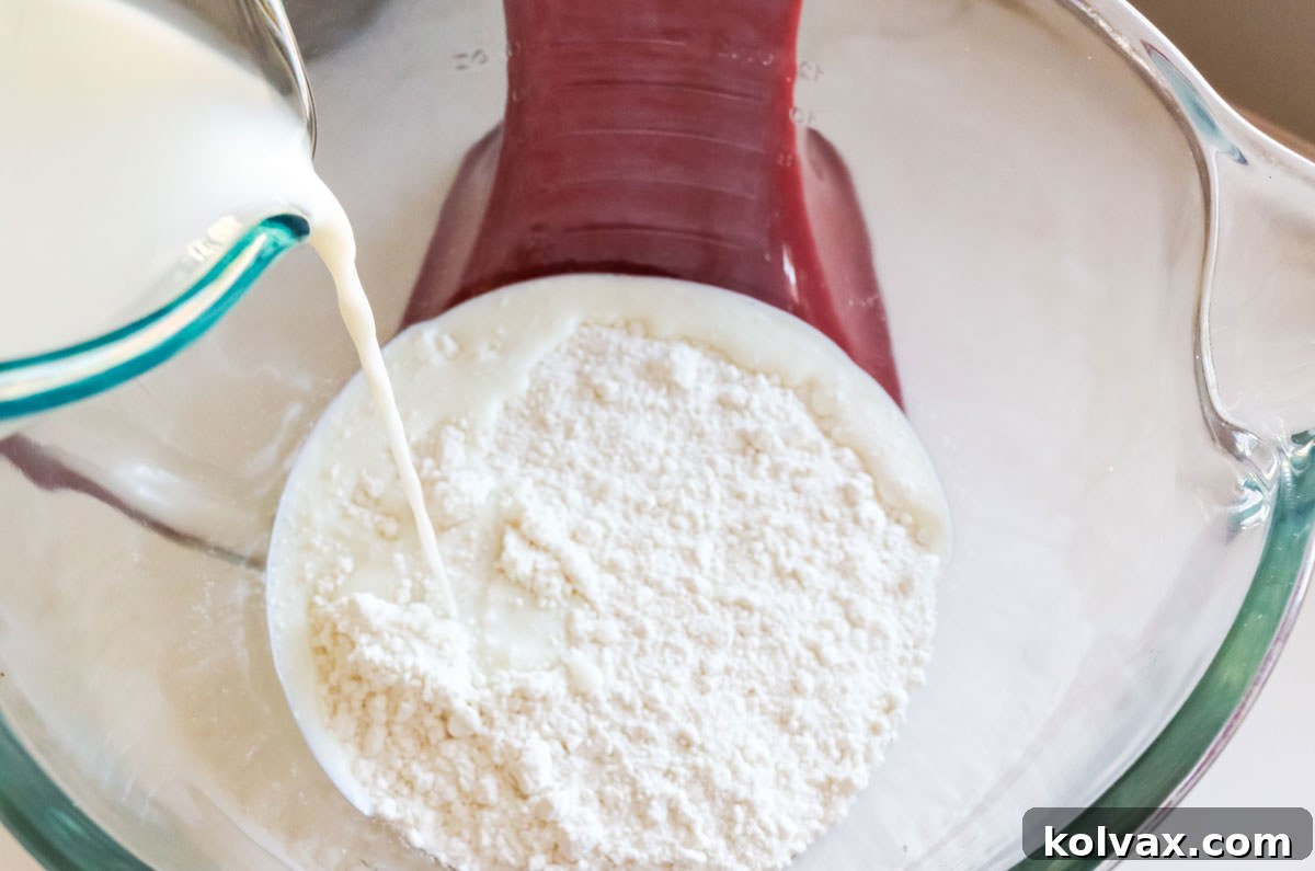 A stream of fresh milk, specifically 2%, being poured into a large mixing bowl already containing dry white cake mix. The focus is on the liquid blending with the powder.