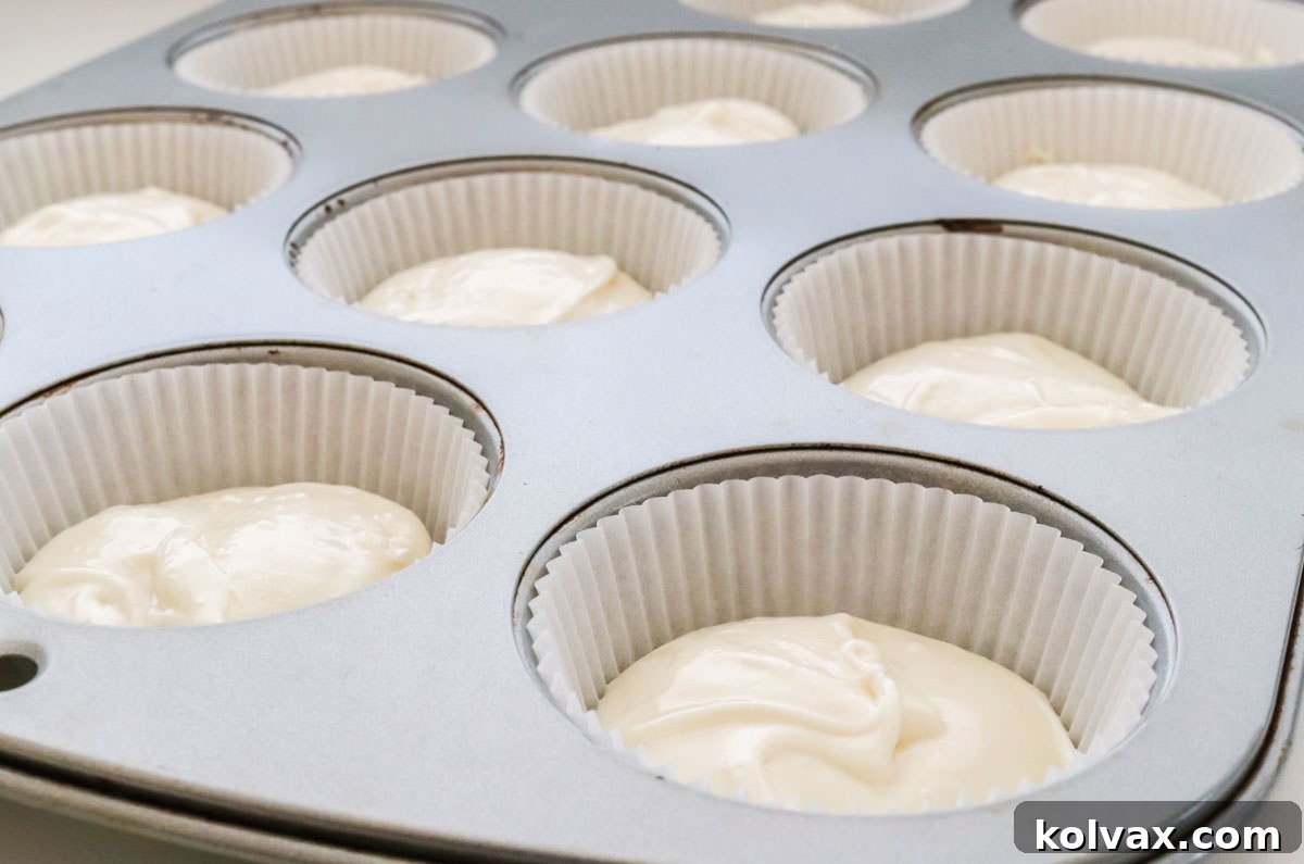 A close-up shot of a cupcake pan with white paper liners, each generously filled with smooth, pure white cake batter, ready for baking. The batter looks perfectly mixed.