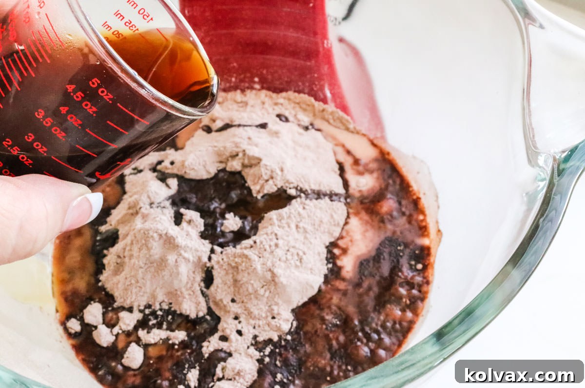 A dark, steaming stream of freshly brewed coffee being poured into a mixing bowl containing chocolate cake batter. The rich liquid mixes with the dark batter.