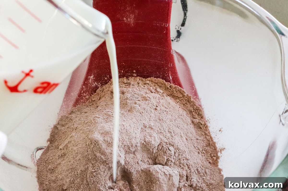 Fresh milk being poured from a measuring cup into a stand mixer's bowl, which already contains chocolate cake mix and other wet ingredients, mid-mixing.
