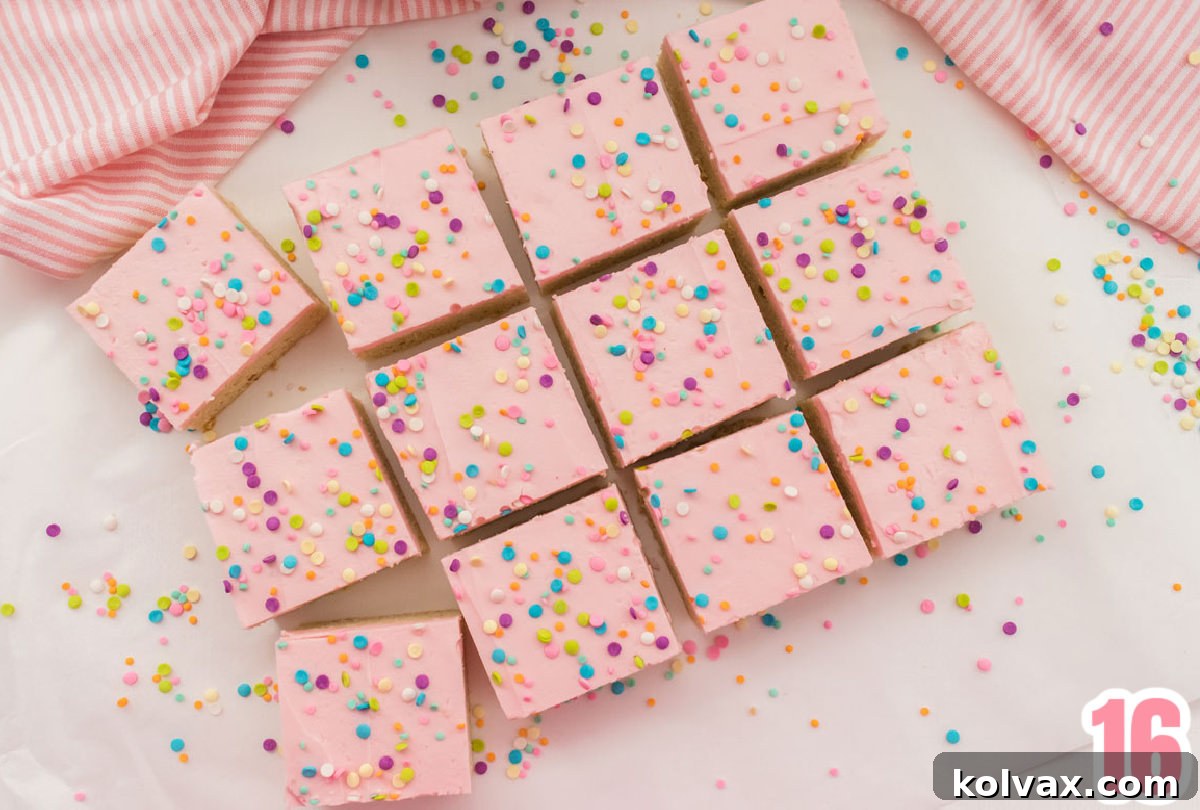 An inviting overhead shot of 12 freshly made Sugar Cookie Bars adorned with pink buttercream frosting and pastel sprinkles, arranged on a pristine white table alongside extra sprinkles and a decorative pink striped kitchen towel.