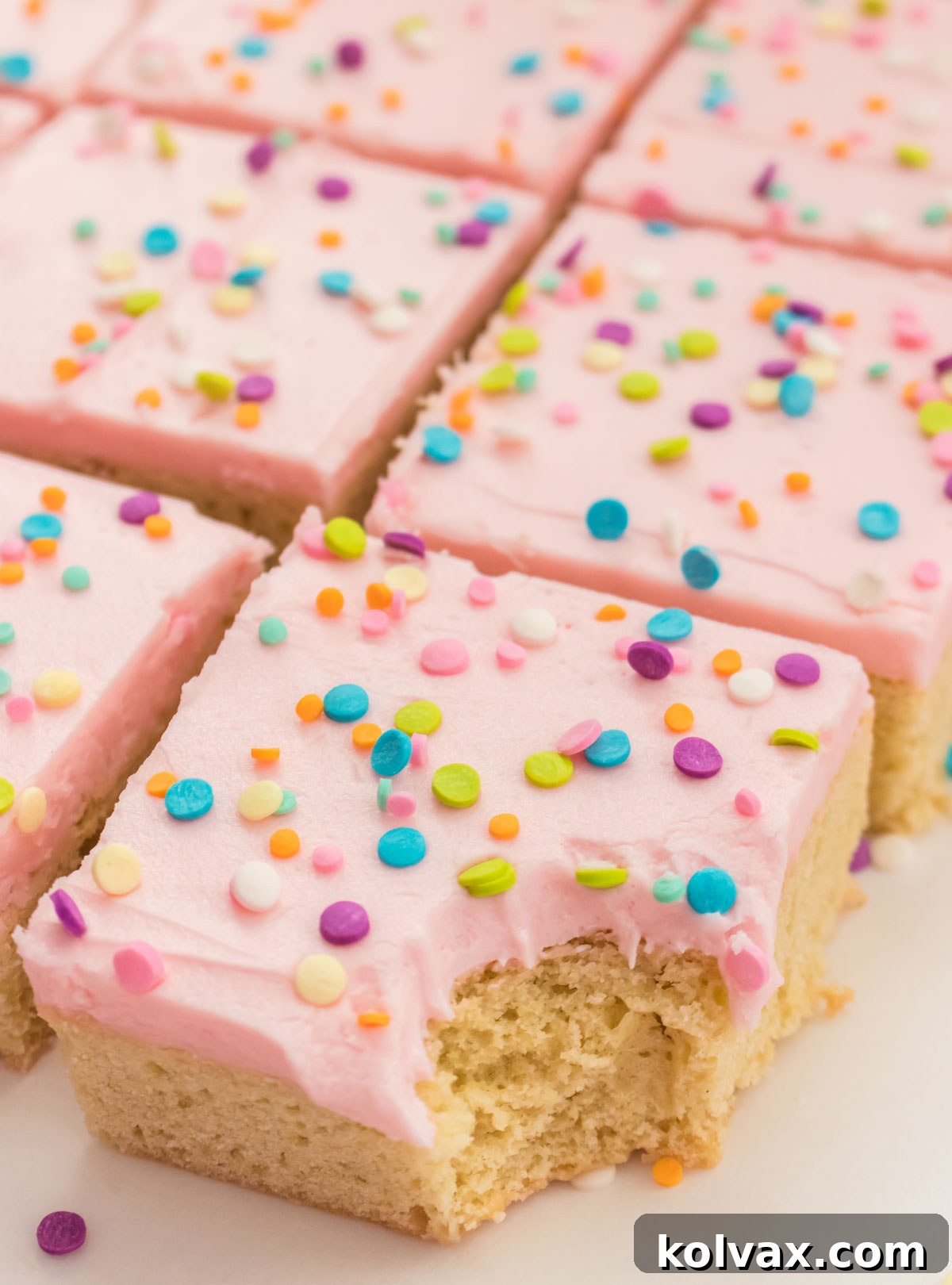 A delicious closeup of rows of Sugar Cookie Bars generously covered in pink buttercream frosting and adorned with pastel sprinkles, with the first bar showing a tempting bite taken out of it.