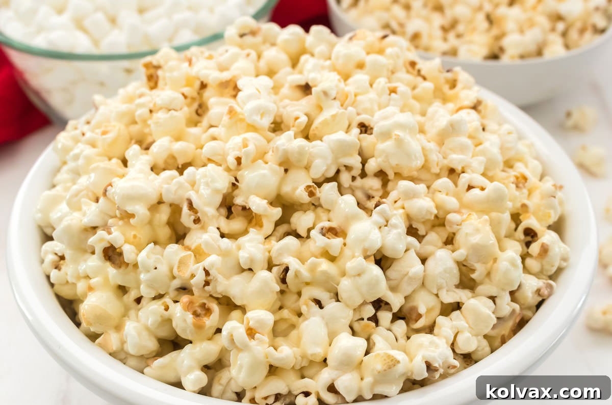 Close up of a white serving bowl filled with Marshmallow Popcorn, with a glass bowl of mini marshmallows and a white bowl of regular popcorn in the soft background.