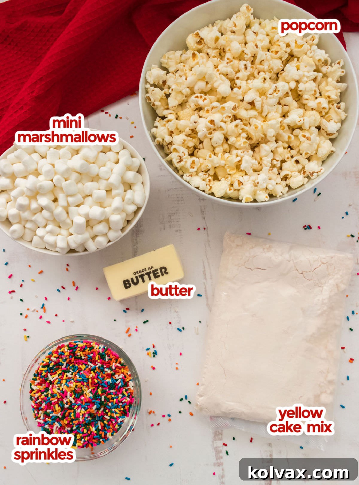 All the essential ingredients neatly displayed for making Cake Batter Popcorn, including fluffy popped popcorn, miniature marshmallows, creamy butter, vibrant rainbow sprinkles, and a box of yellow cake mix.