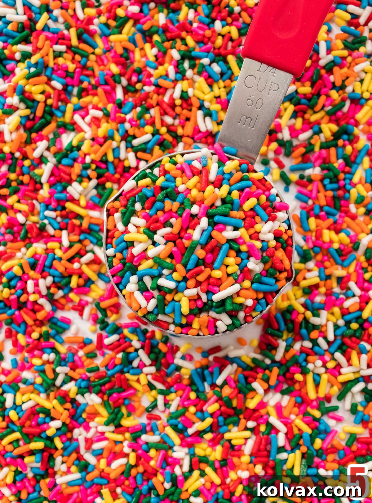 Closeup on a scattering of vibrant rainbow sprinkles spread across a white surface, with a 1/4 measuring cup full of sprinkles positioned centrally.