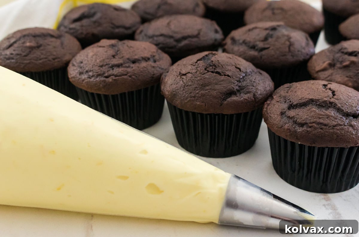 A decorator bag filled with Lemon Cream Cheese Icing sitting on a white table in front of a batch of unfrosted chocolate cupcakes.