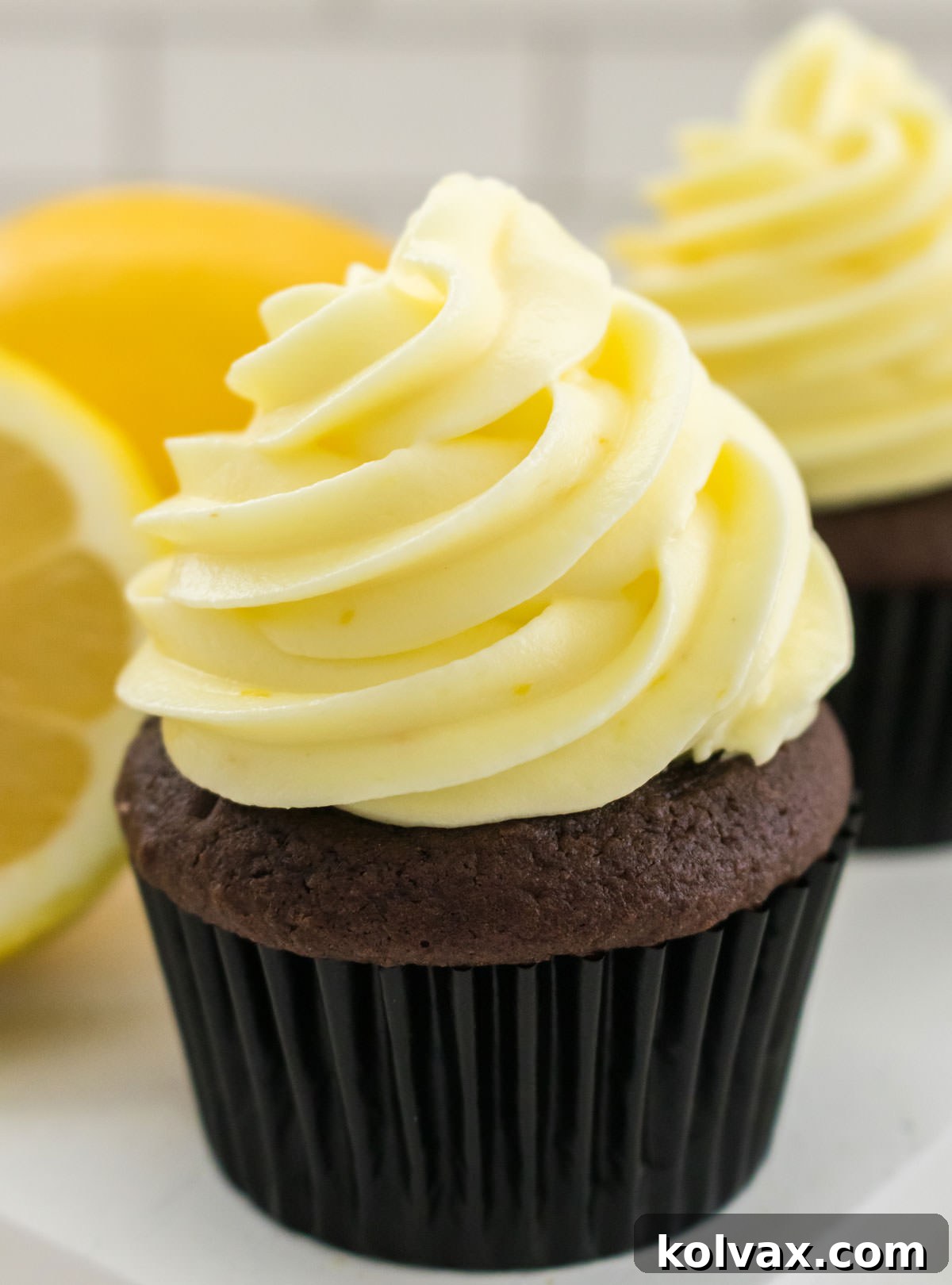Closeup of a Chocolate Cupcake topped with a swirl of The Best Lemon Cream Cheese Frosting sitting on a white plate in front of a fresh lemon.