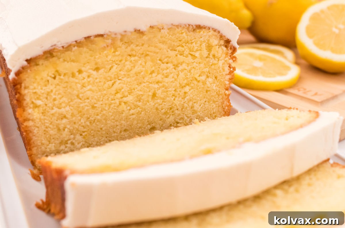 Close-up of a perfectly frosted Lemon Pound Cake with creamy vanilla icing, sitting on a white plate alongside a wooden cutting board with fresh lemon slices, highlighting its vibrant yellow hue and appealing texture.