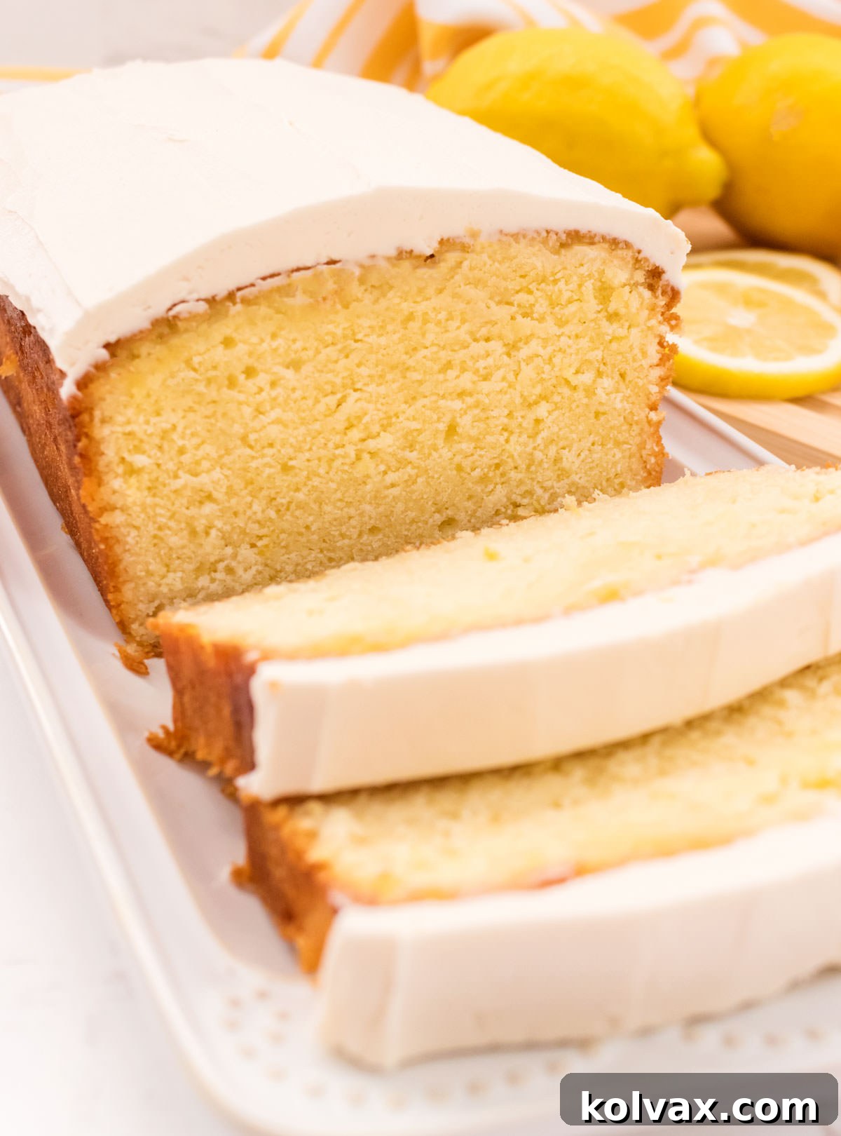 A stunning close-up of a partially sliced Lemon Pound Cake, artfully arranged on a white serving platter on a pristine white table, with blurred fresh lemons in the background, ready to be enjoyed.