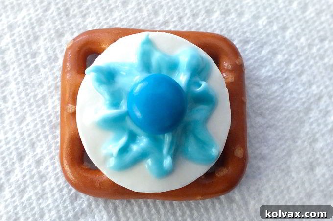 Adding Blue M&M to the Snowflake Pretzel Bites - The final touch for your Disney Frozen Snowflake Pretzel Bites: carefully placing a blue M&M Mini in the center of the crafted light blue snowflake pattern, ensuring the logo faces down for a clean look.