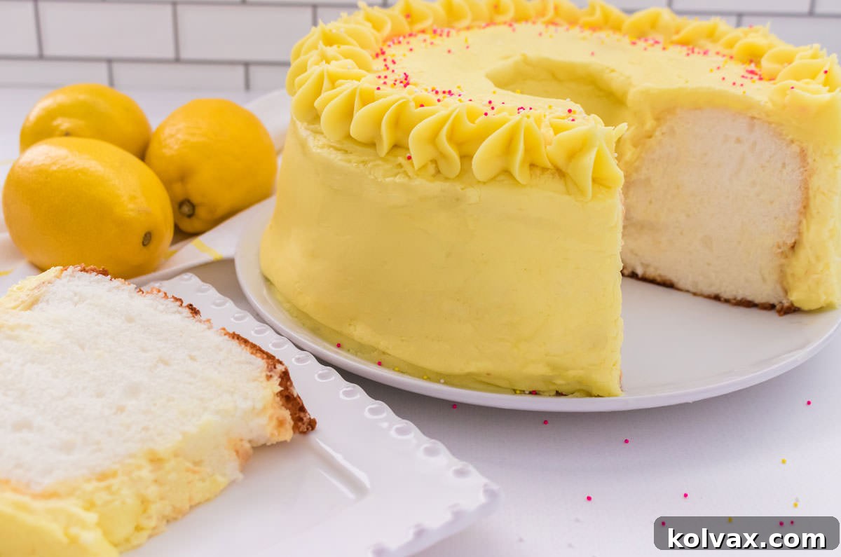 Closeup of an Angel Food Cake with Lemon Frosting on a cake plate on a white table with a slice taken out and on a dessert plate, garnished with lemon zest.