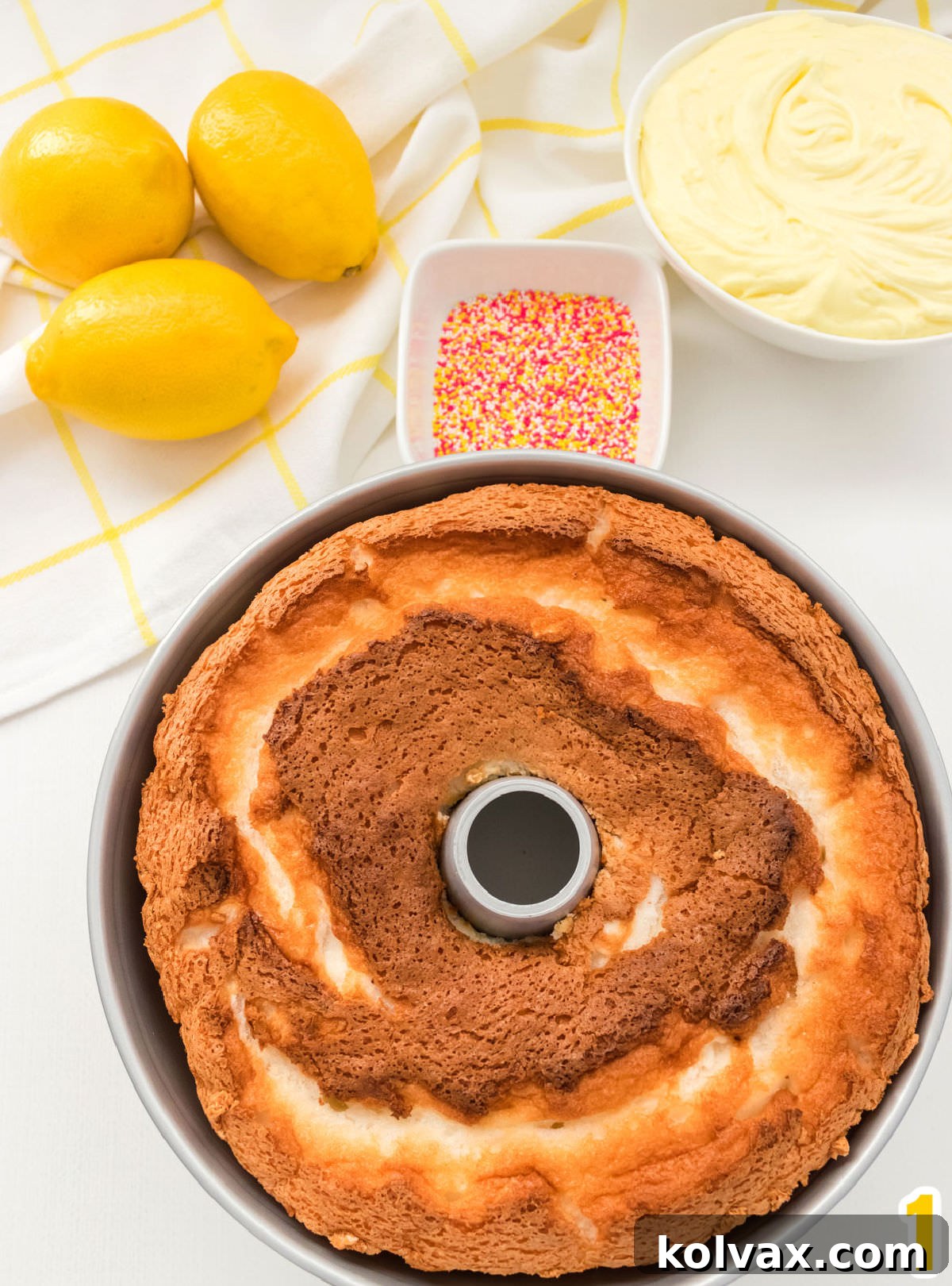 Closeup of an Angel Food Cake just out of the oven and still in the cake pan sitting on a white surface in front of a ramekin of sprinkles and a bowl of lemon frosting.