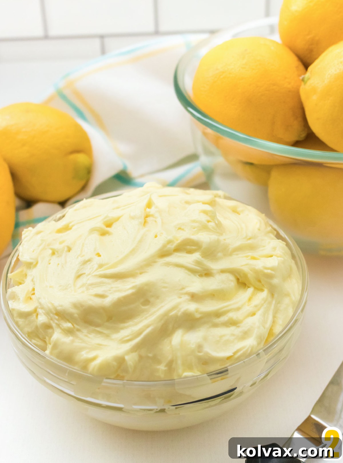 A glass bowl filled with Lemon Frosting sitting in front of a glass bowl filled with Lemons.
