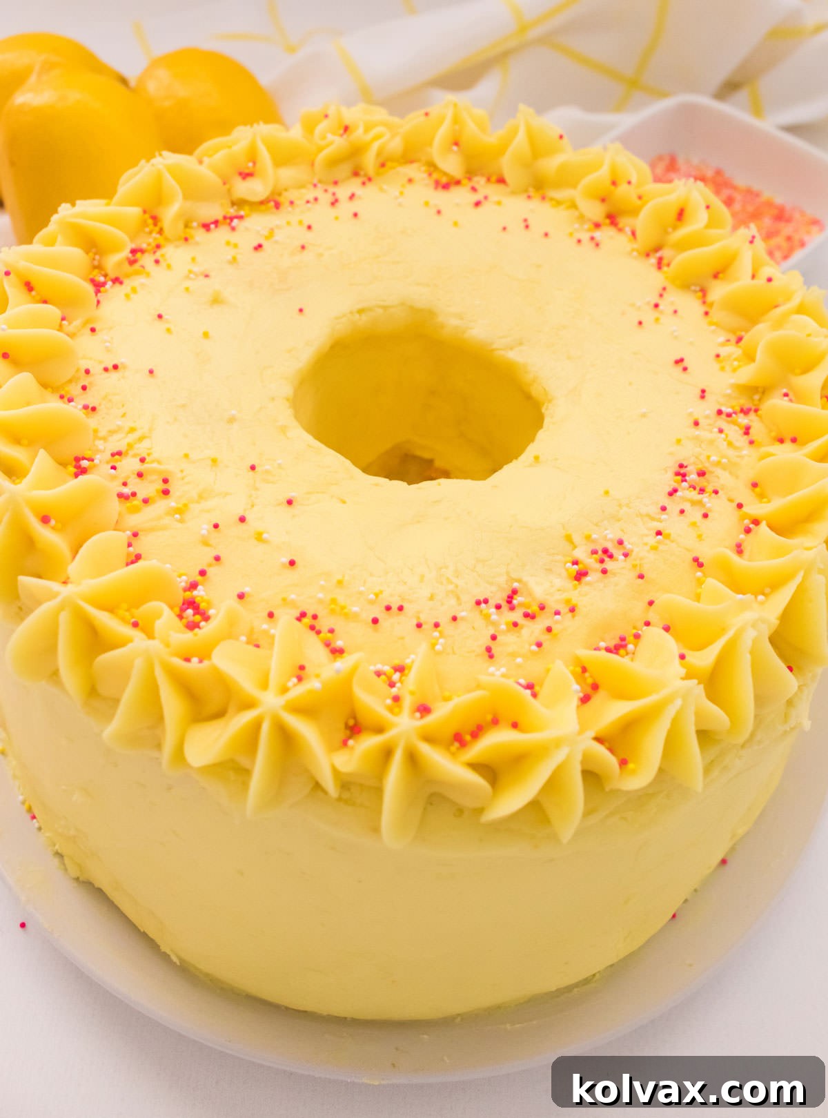 Closeup on an Angel Food Cake with Lemon Frosting sitting on a white cake plate on a white table with lemons in the background.