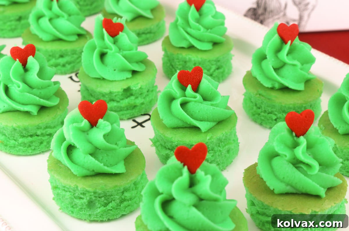 A close-up view of a white platter laden with vibrantly green Grinch Cake Bites, each topped with a swirl of green frosting and a single red heart sprinkle, ready for a festive Christmas gathering.