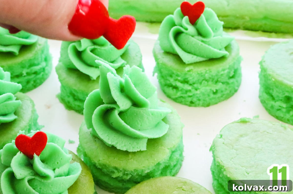A hand gently placing a vibrant red heart sprinkle onto the freshly piped green frosting swirl of a Grinch Cake Bite, completing the festive look.