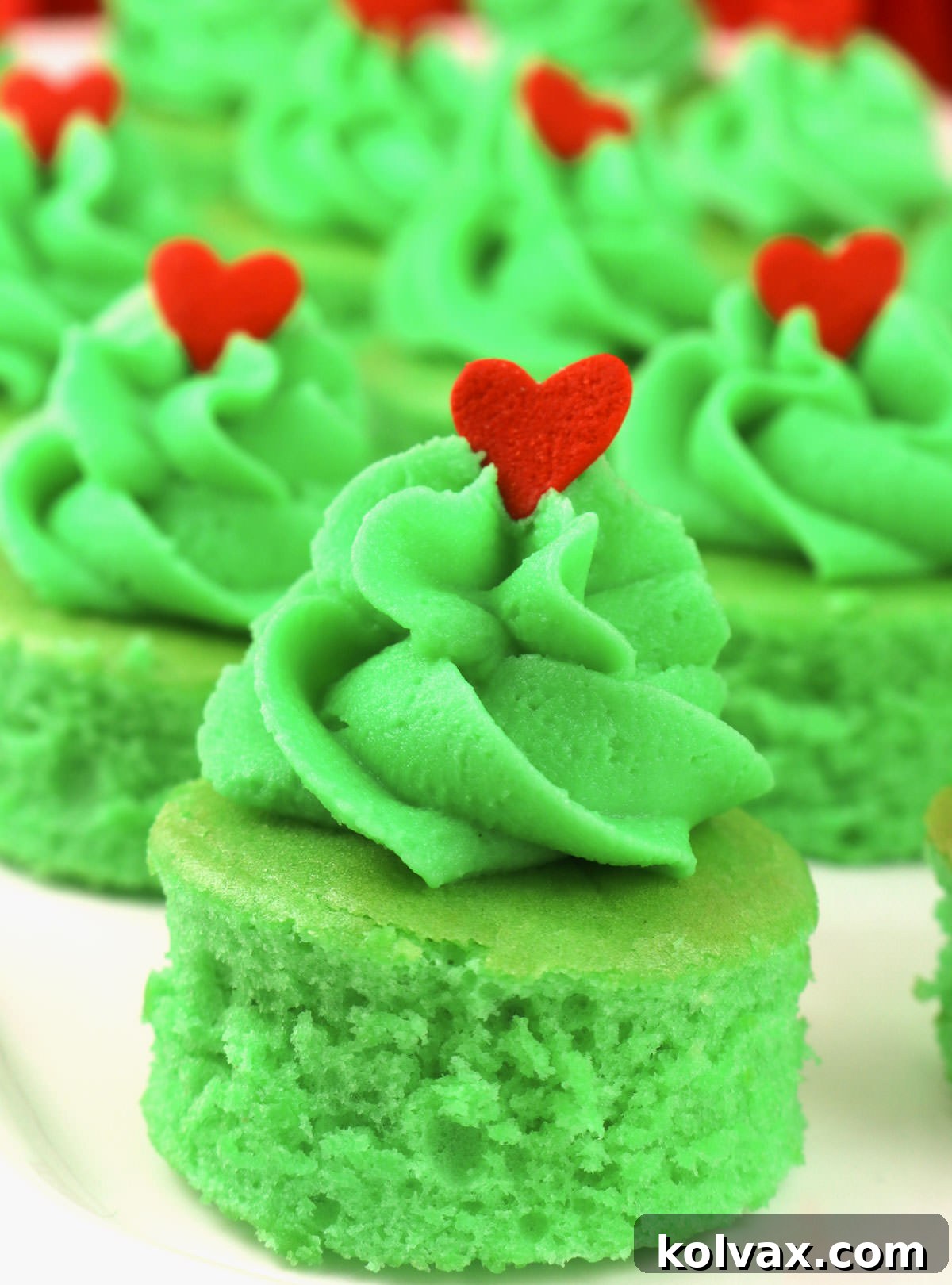A pristine white serving platter holds a generous batch of finished Grinch Cake Bites, each adorned with green frosting and a red heart, ready to be enjoyed at a holiday party.