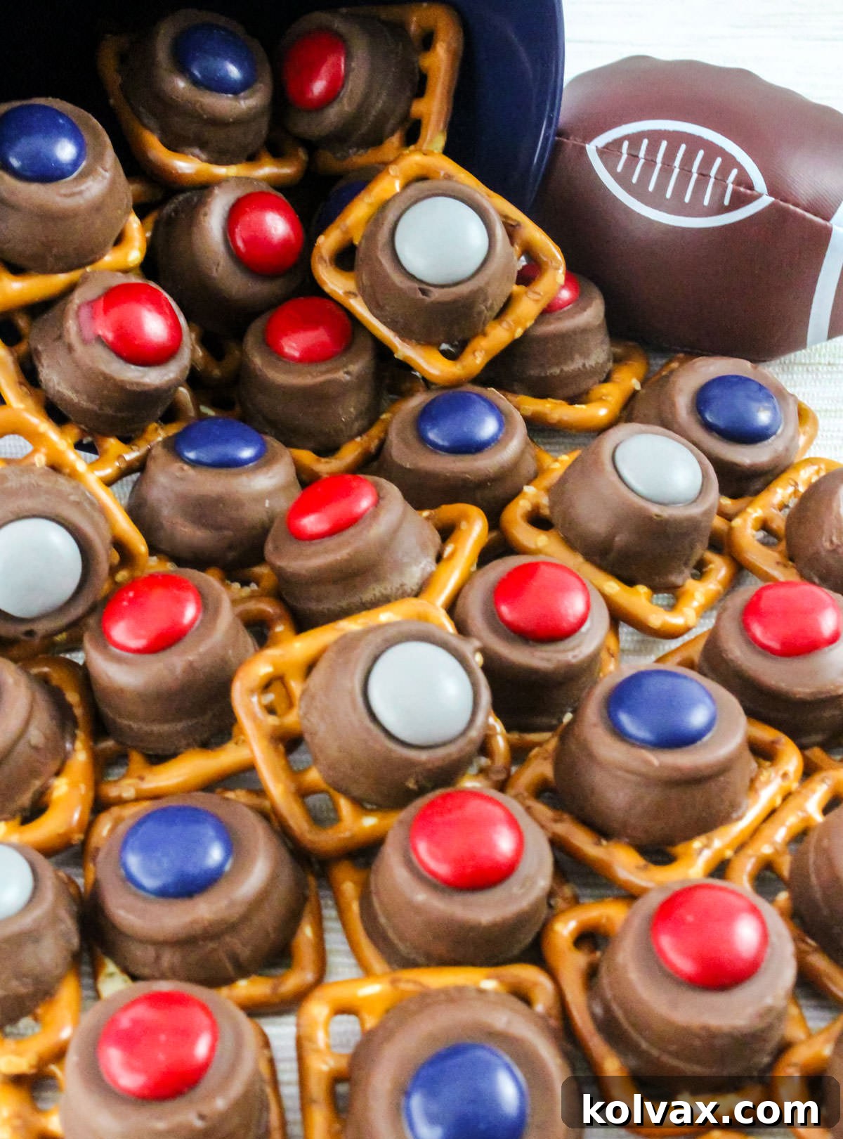 A generous quantity of colorful New England Patriots Pretzel Bites, featuring mini pretzels with red, blue, and silver candy melts, overflowing from a blue container next to a small decorative football.