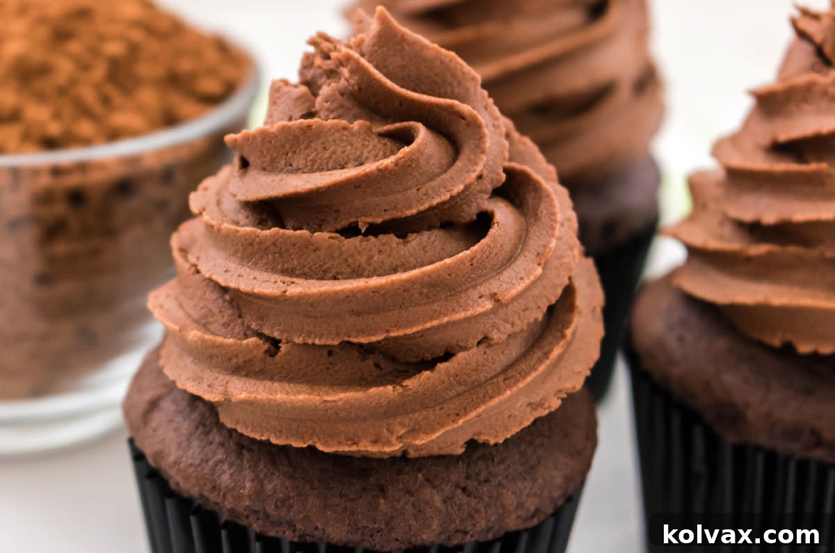 Three decadent chocolate cupcakes adorned with generous swirls of our signature Chocolate Mint Buttercream Frosting, elegantly presented on a crisp white surface next to a small glass bowl brimming with unsweetened cocoa powder, highlighting the key ingredient.