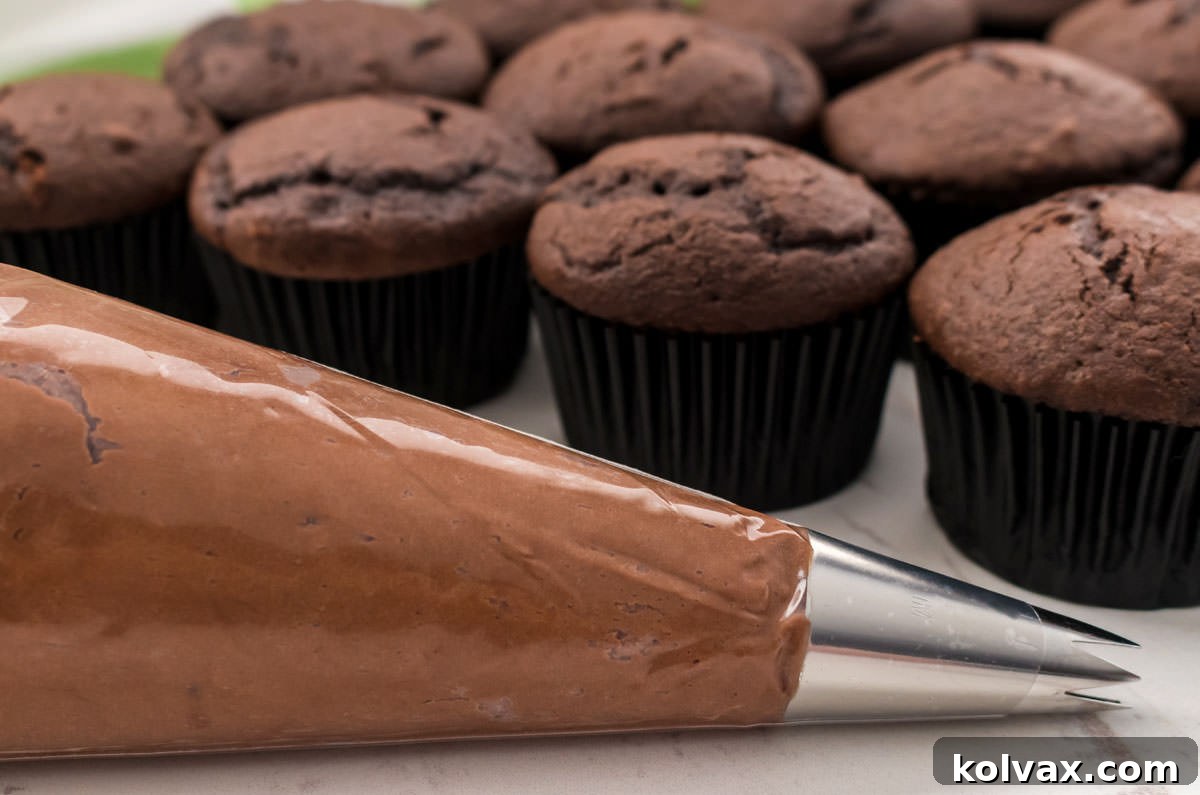 A piping bag generously filled with the vibrant green Best Chocolate Mint Buttercream Frosting, poised on a pristine white surface, with a batch of unfrosted chocolate cupcakes waiting in the background, ready for their delicious topping.